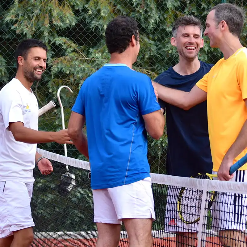 Four men standing on a tennis court near the net, smiling and interacting with tennis rackets in hand.