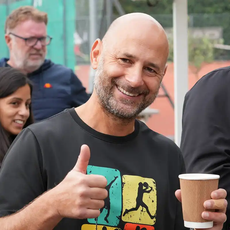 Smiling bald man giving a thumbs-up with one hand and holding a coffee cup in the other at an outdoor gathering.