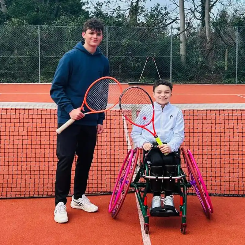 Two young men on a tennis court, one standing holding a tennis racket and the other seated in a wheelchair with a tennis racket, smiling at the camera.
