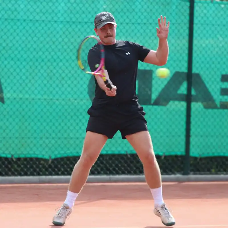 Man in black sportswear and cap hitting a tennis ball with a racket on a clay court.