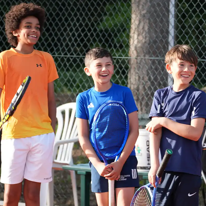Young blond boy wearing navy shirt and green shorts preparing to hit a tennis ball with a racket on an outdoor court.