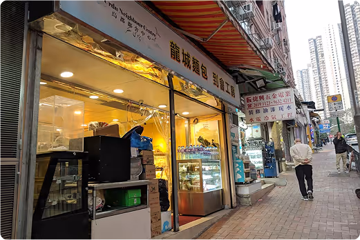 Street view of a Hong Kong neighborhood showing Bread Bunch, UNC's affiliate, including a brightly lit bakery or food store, with high-rise residential buildings in the background