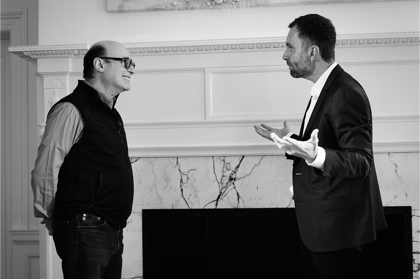 Two men engaged in conversation while standing in front of a cozy fireplace.
