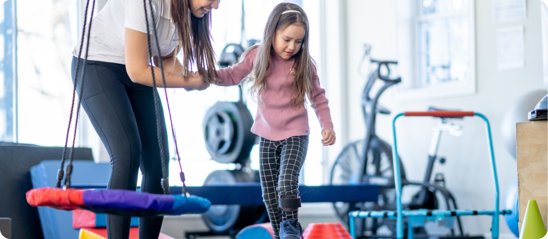 Erwachsene Person unterstützt ein Kind beim Balancieren über einen Parcours in einem Raum mit Trainingsgeräten