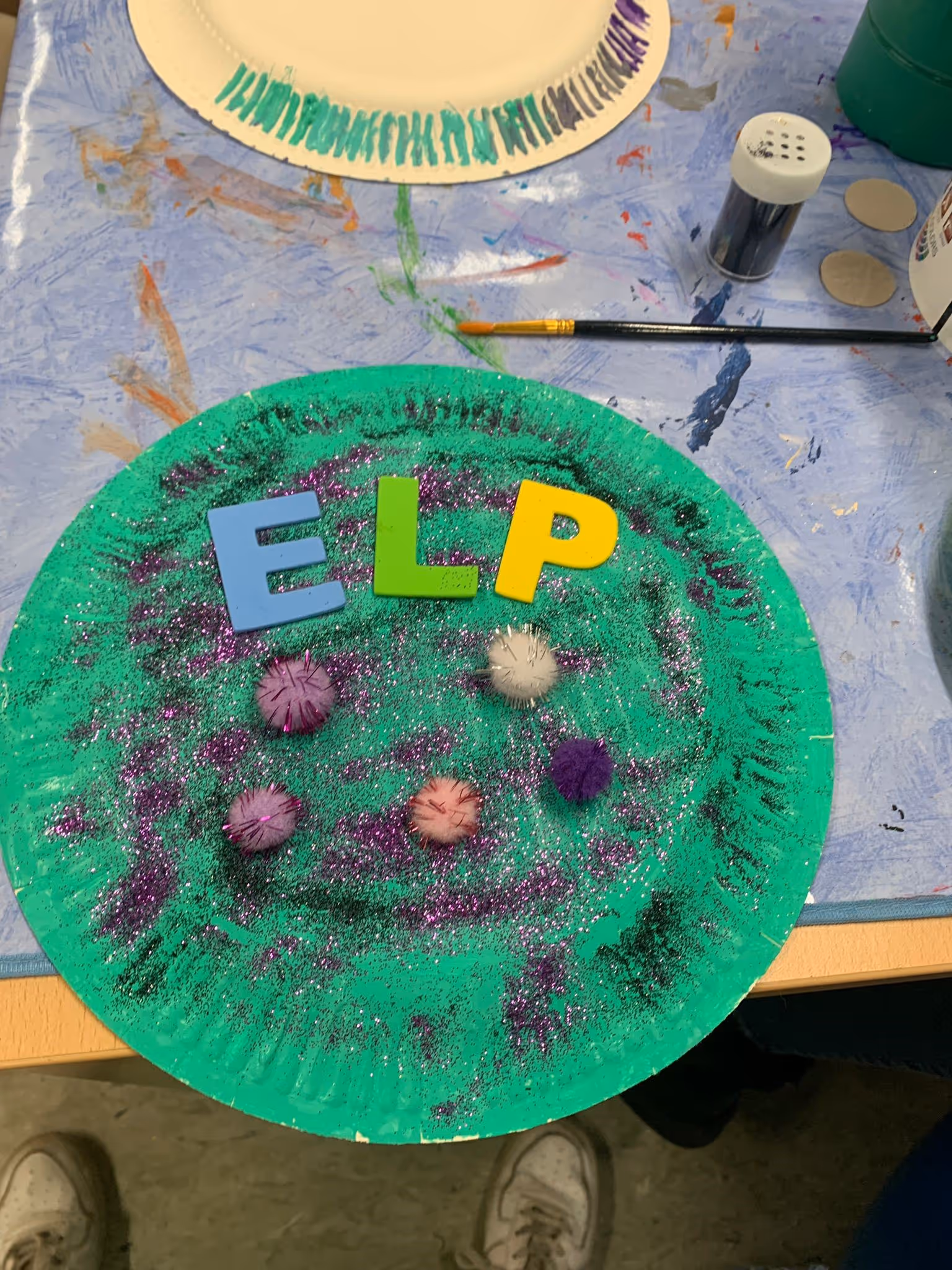 Grüner bemalter Pappteller mit bunten Buchstaben und aufgeklebten Naturmaterialien auf einem Basteltisch