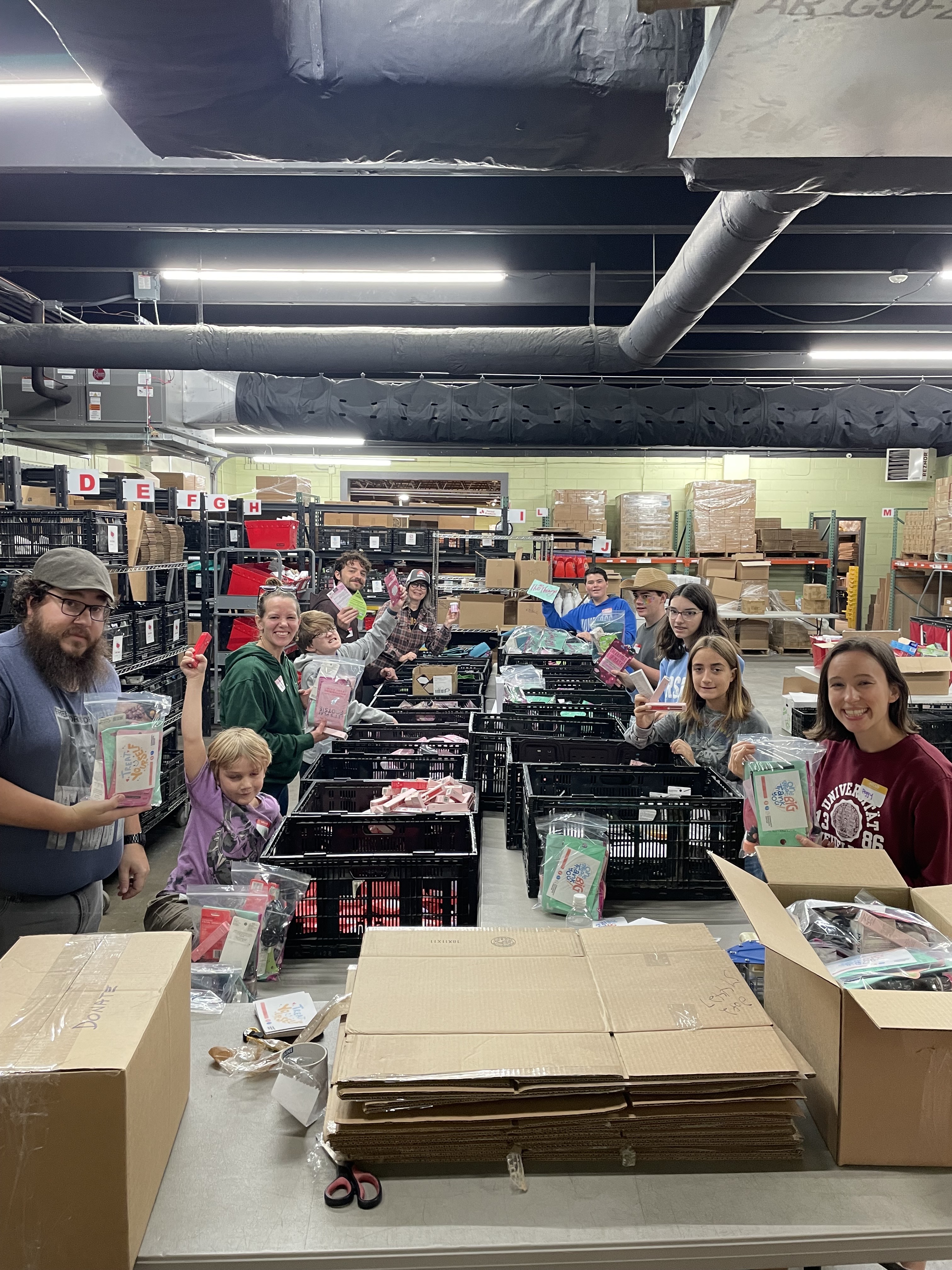 Group of volunteers standing around tables with boxes and black crates filled with packaged supplies in a warehouse setting.