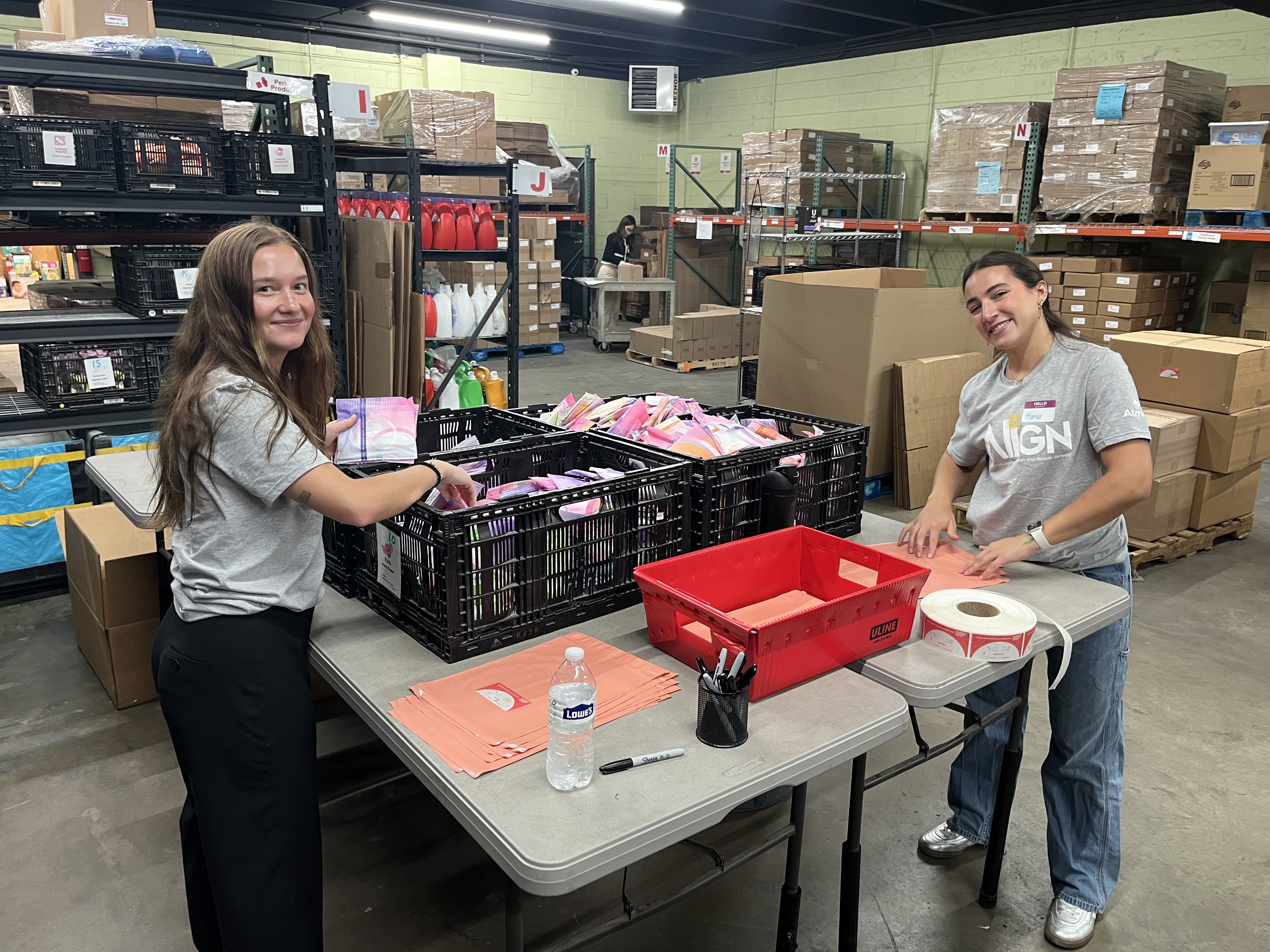 Two women smiling and organizing items on a table in a warehouse filled with boxes and shelves.