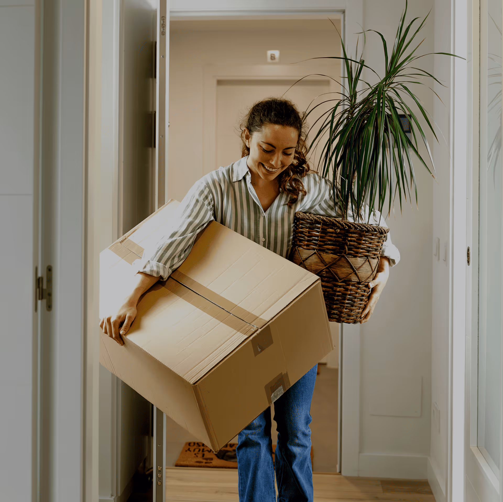 Mulher sorridente a entrar em casa com uma caixa de cartão e uma planta numa cesta de vime.