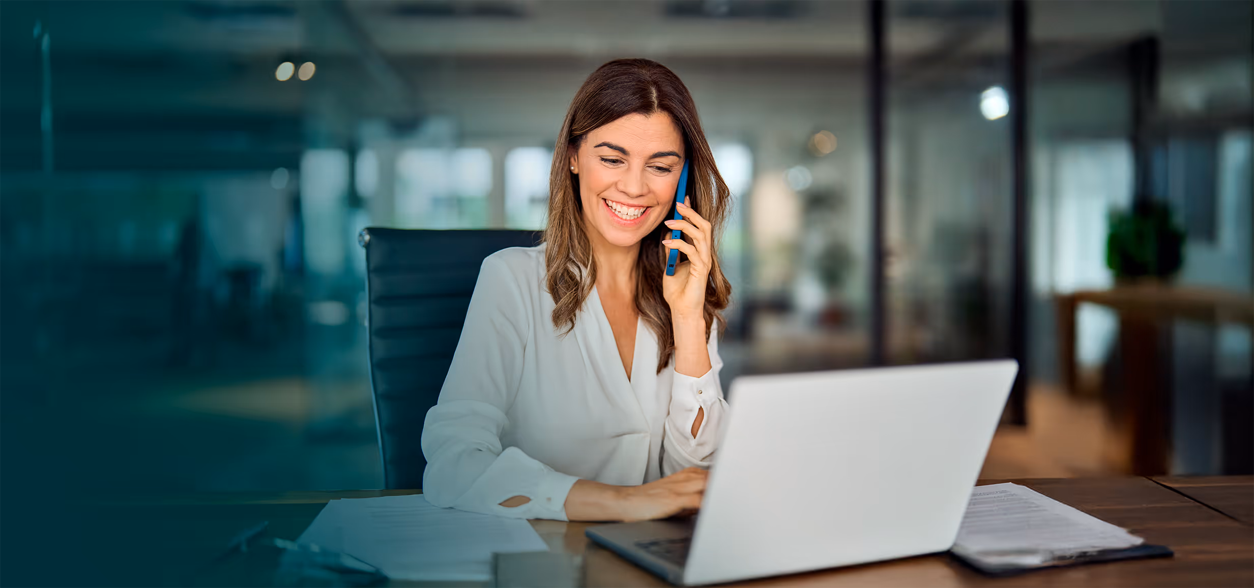 Mulher sorridente a falar ao telemóvel enquanto trabalha num computador portátil num escritório moderno.