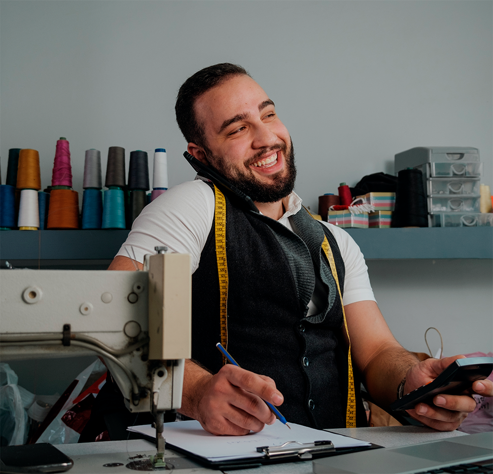 Costureiro sorridente a falar ao telemóvel segurando uma calculadora e escrevendo num bloco de notas, com fita métrica pendurada no pescoço.