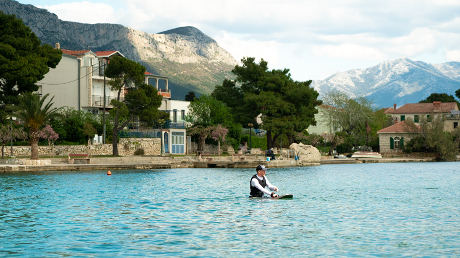 Man kneeling on a electric surfboard in calm water with houses, trees, and mountains in the background under a partly cloudy sky.