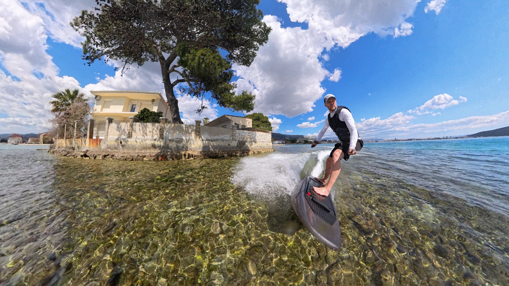 Man riding a motorized surfboard on clear shallow water near a stone-walled house and tree under a partly cloudy blue sky.