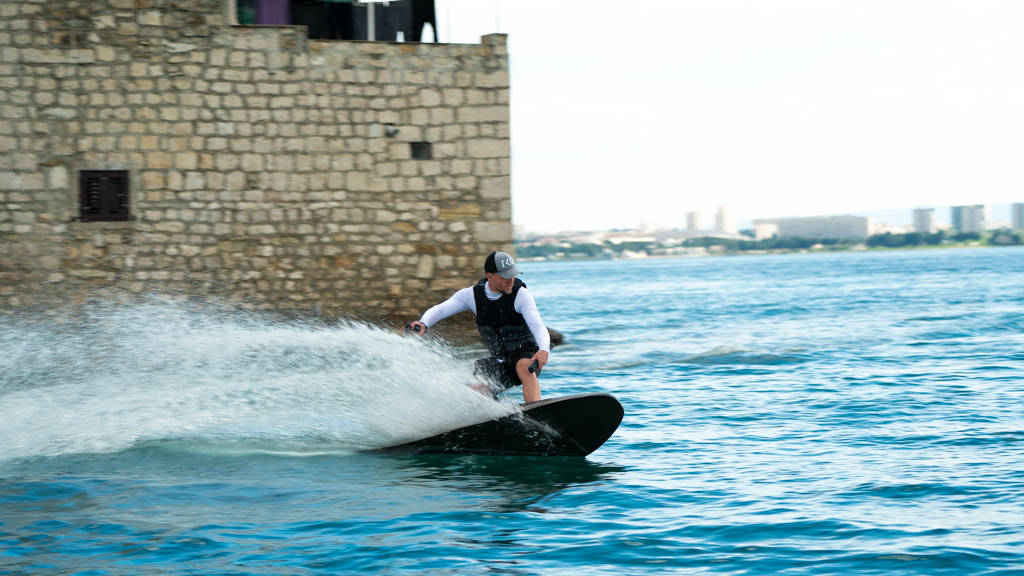 Man in life vest and cap riding a black electric surfboard, creating a spray of water near stone wall by the sea.