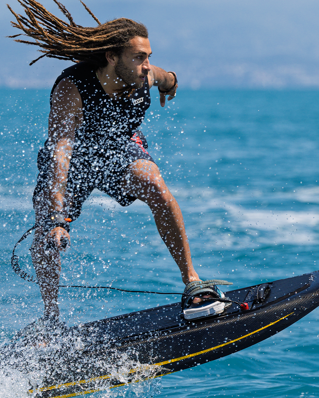 Man with long dreadlocks rides a jet surfboard on blue water, splashing water around him.