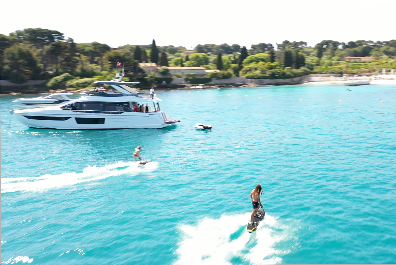 Two people riding electric surfboards on clear blue water near a white luxury yacht with green trees in the background.