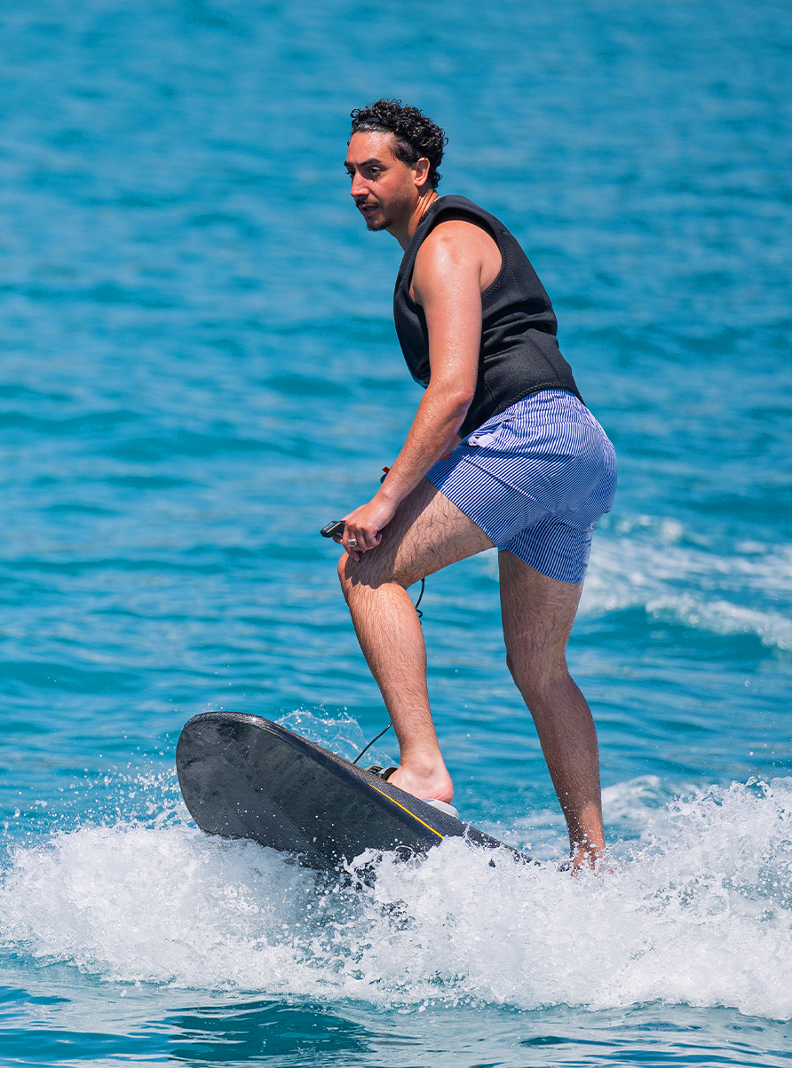 Man in black life vest and blue striped shorts riding an electric surfboard on the ocean.