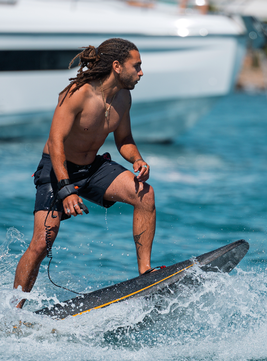 Shirtless man with dreadlocks riding an electric surfboard on the water near a white boat.