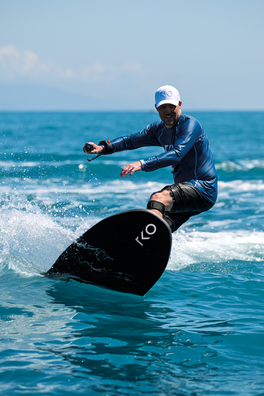 Man in blue shirt and white cap riding a black jetboard on ocean water.