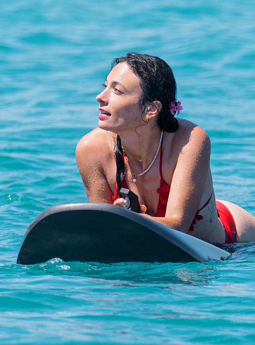 Woman in a red bikini lying on a jetboard in the water, looking to the side with a flower in her hair.