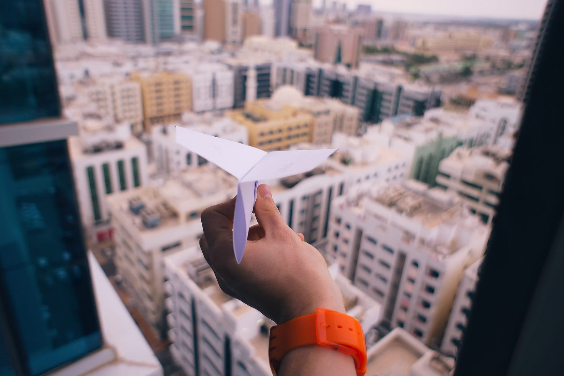 A hand with an orange wristband holds a white paper airplane, overlooking a blurred cityscape from a high window.
