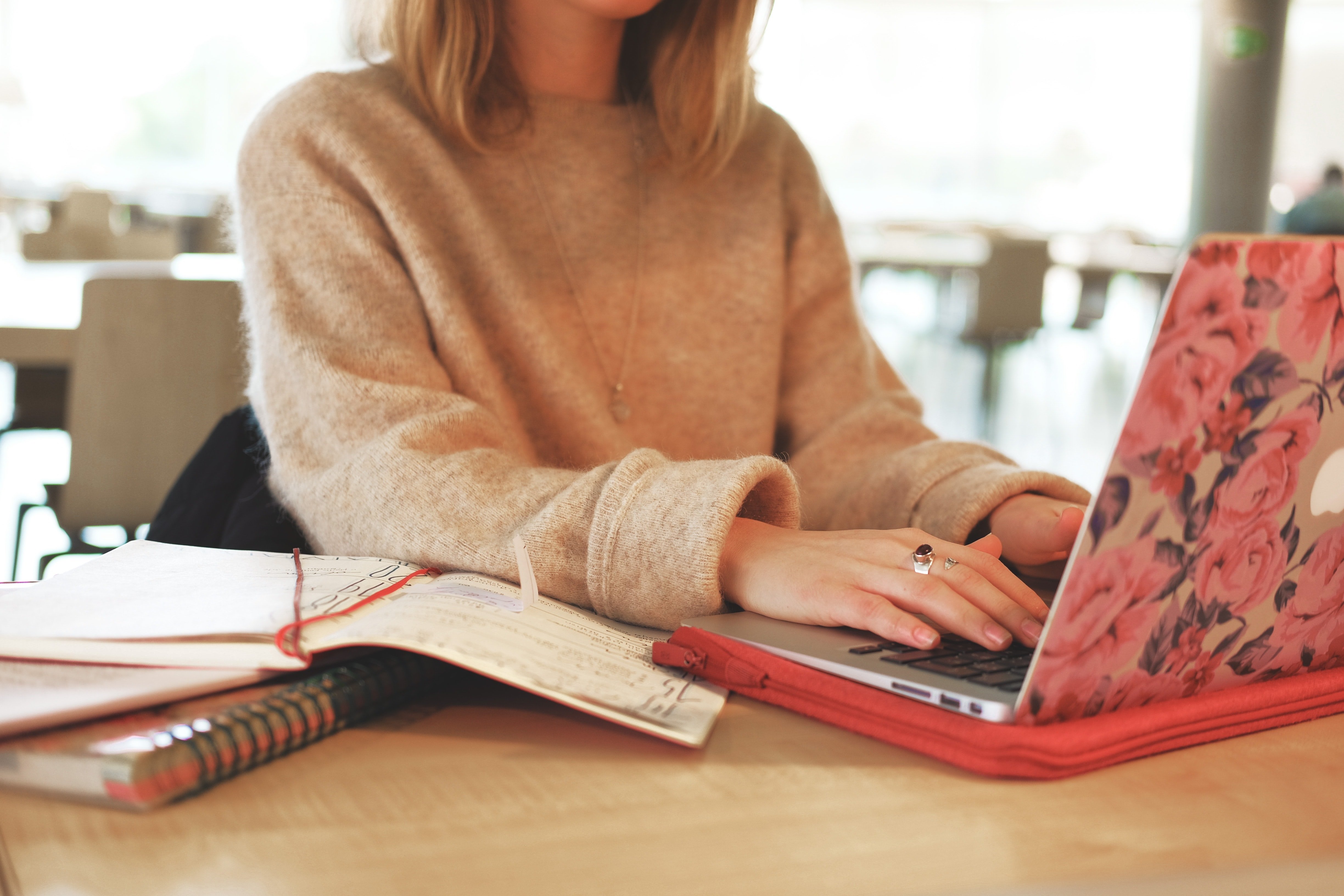 Person in brown sweater with rings types on floral-lidded laptop at a wooden table with notebooks.