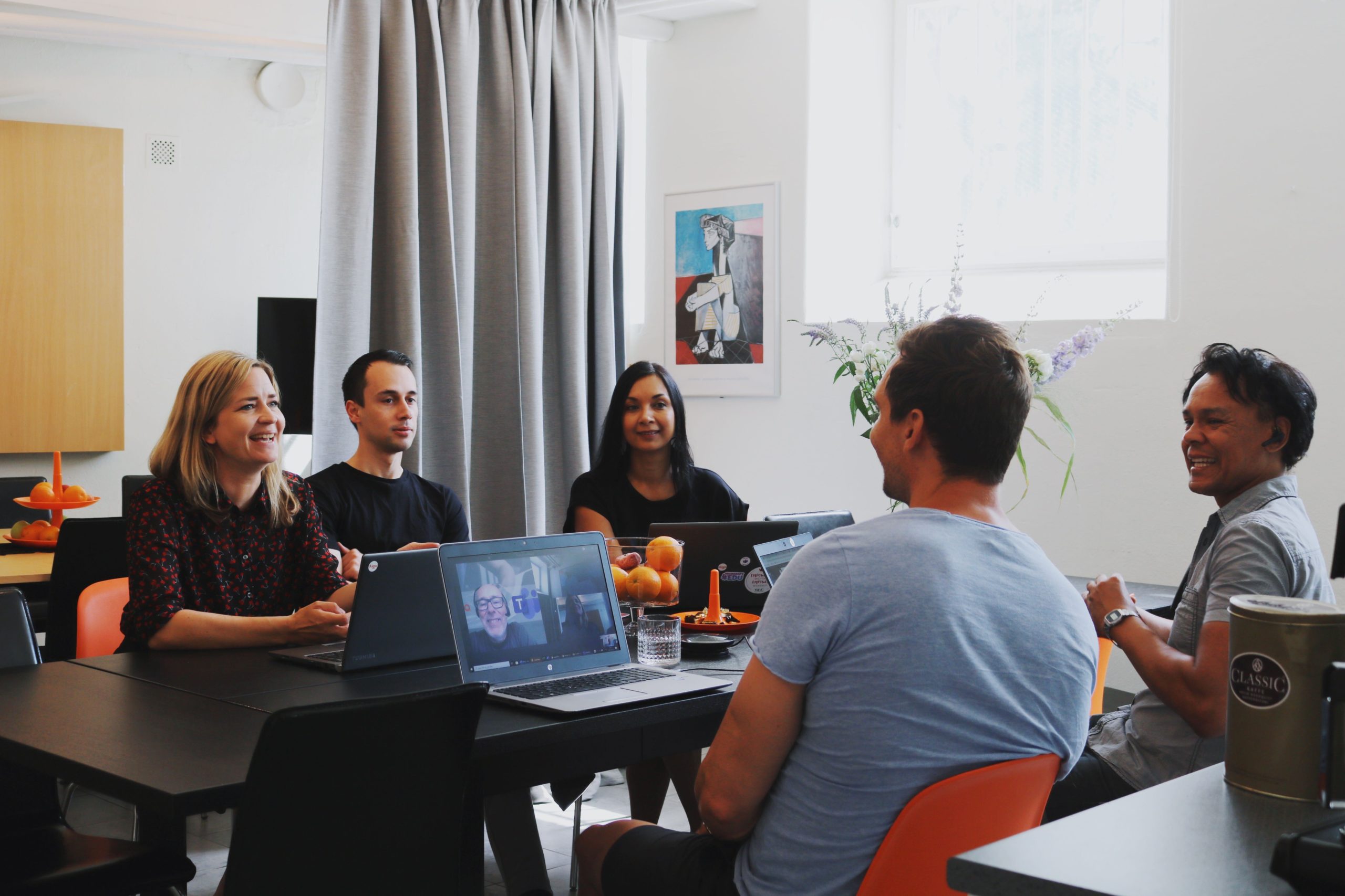 Five people meeting at a table with laptops in a bright room; one shows a video call.