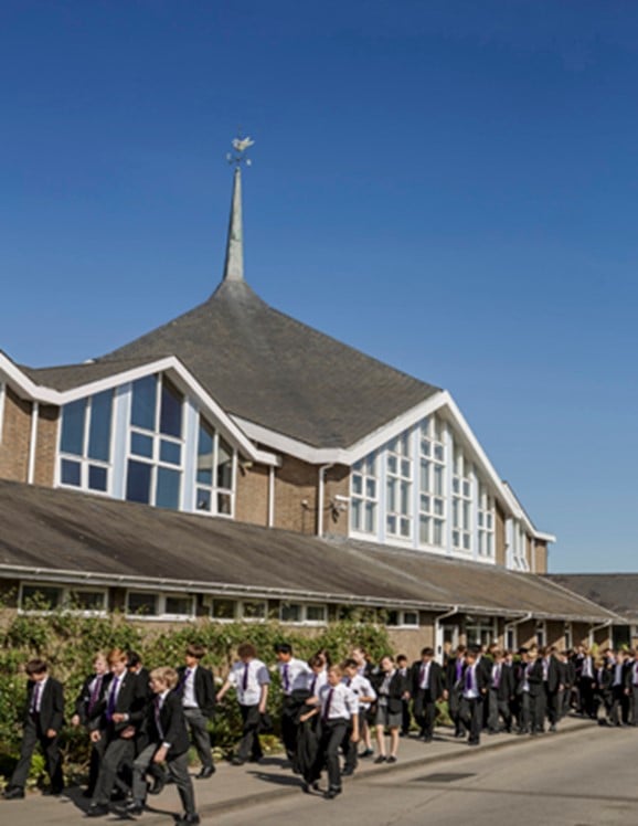 School building with a spire and dove weather vane, a group of students in uniform walk past on a sunny day.