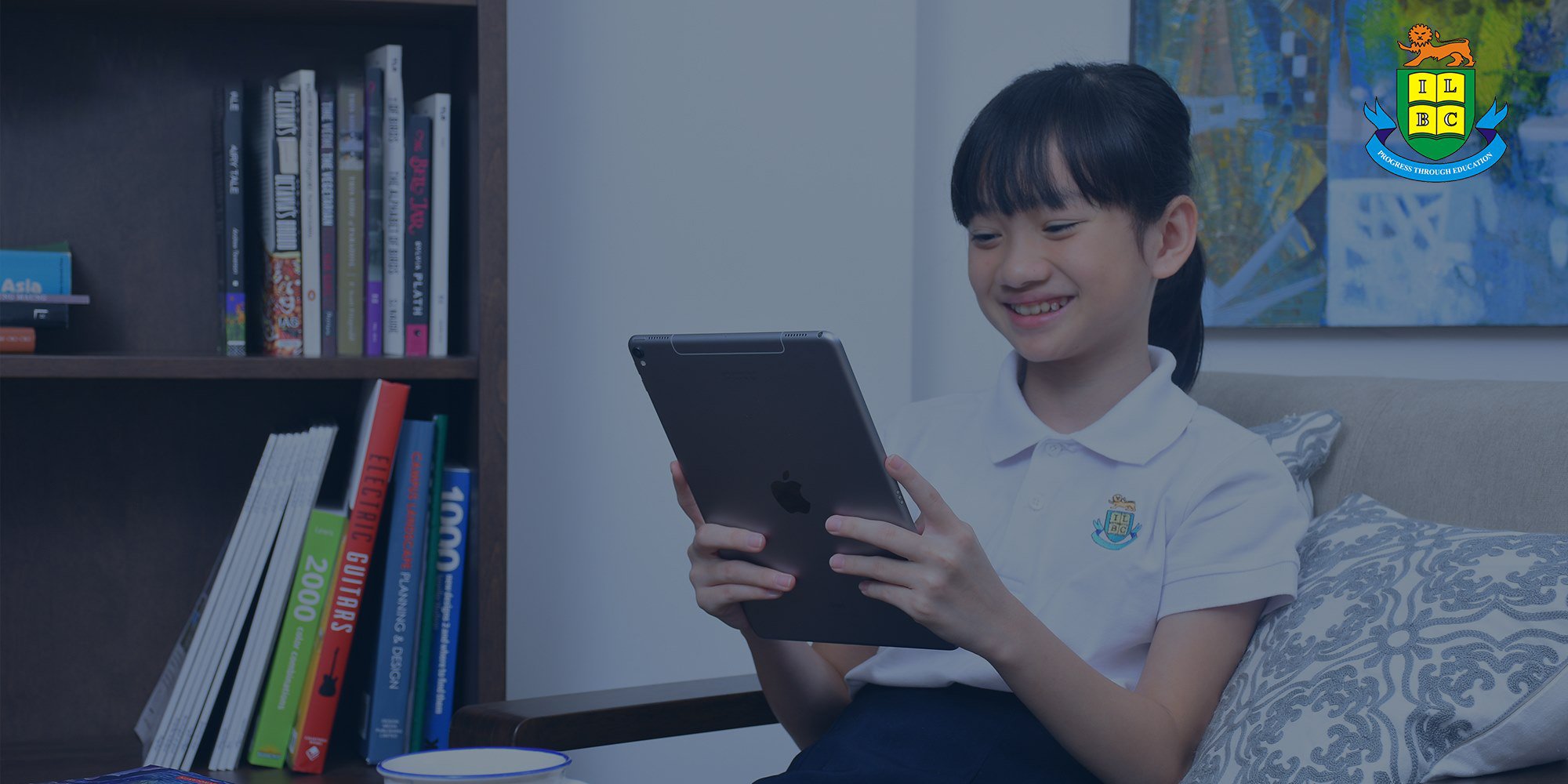 Smiling girl in school uniform holds tablet. Bookshelf with books, and school crest (lion, 'ILBC') in background.