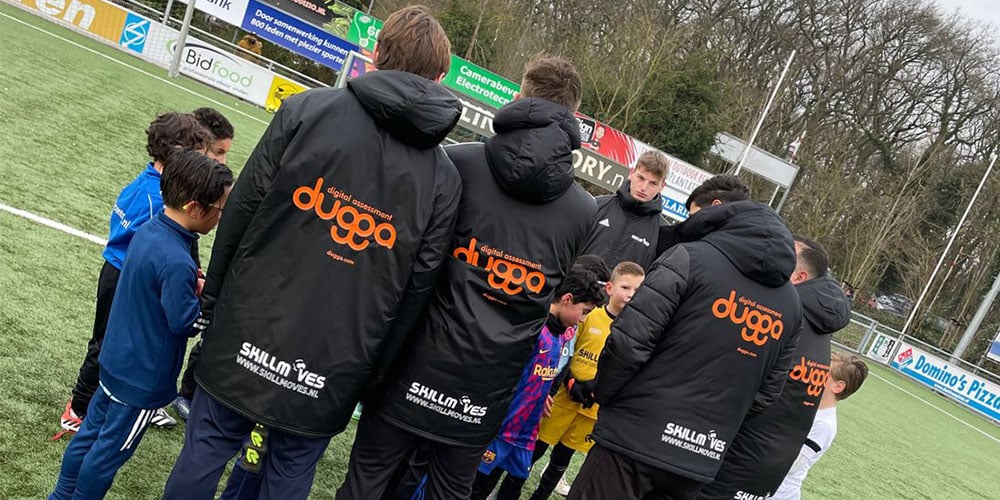 Young soccer players and coaches on a field. Coaches wear jackets with 'duggaa' & 'SKILLMOVES' logos.