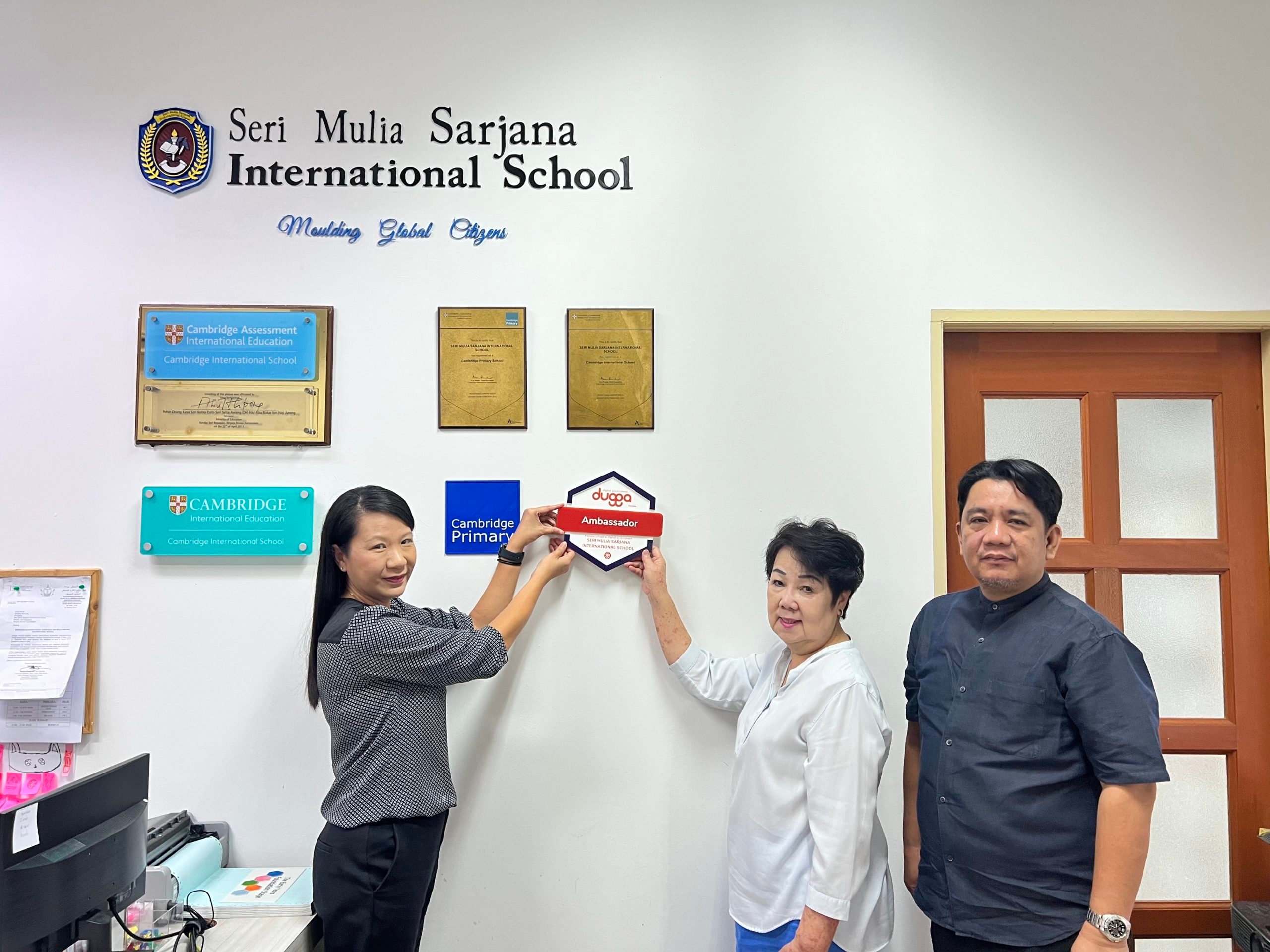 Two women and a man at Seri Mulia Sarjana International School. Women hold "Cambridge Primary" and "Dugga
