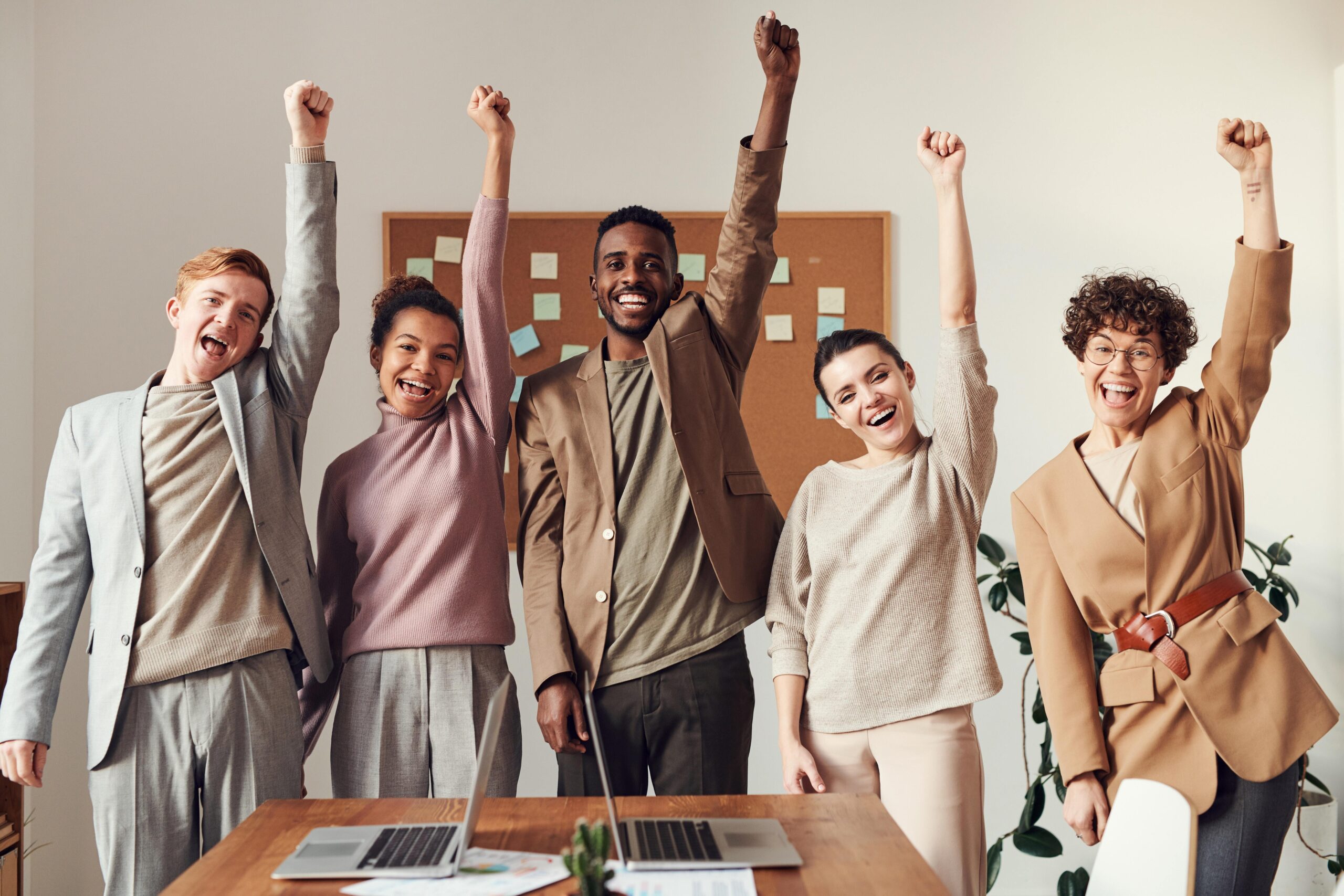 Five diverse people in an office raise fists, smiling broadly, celebrating behind a desk.