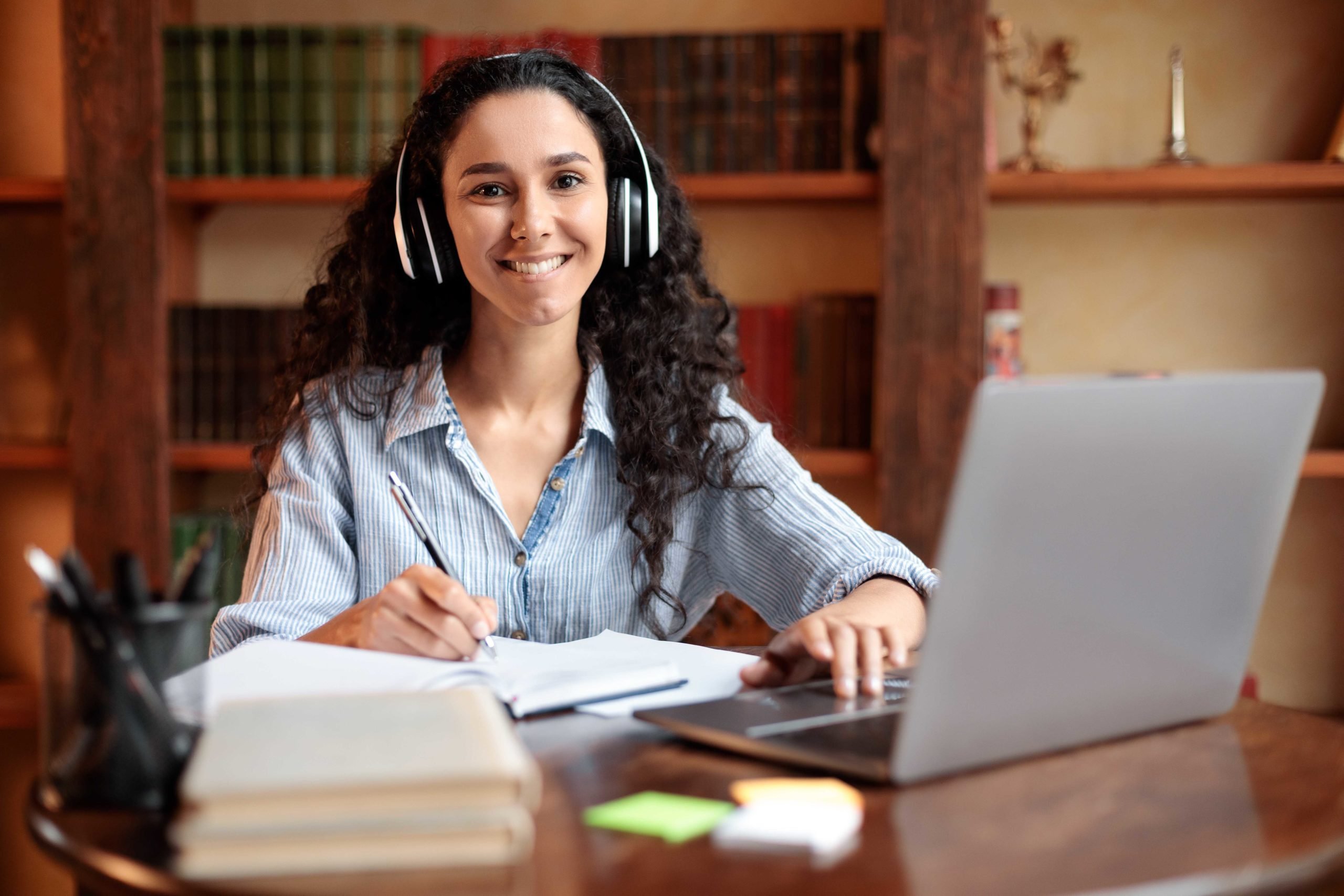 Smiling woman in headphones writes in a notebook, uses laptop at desk with bookshelves in background.