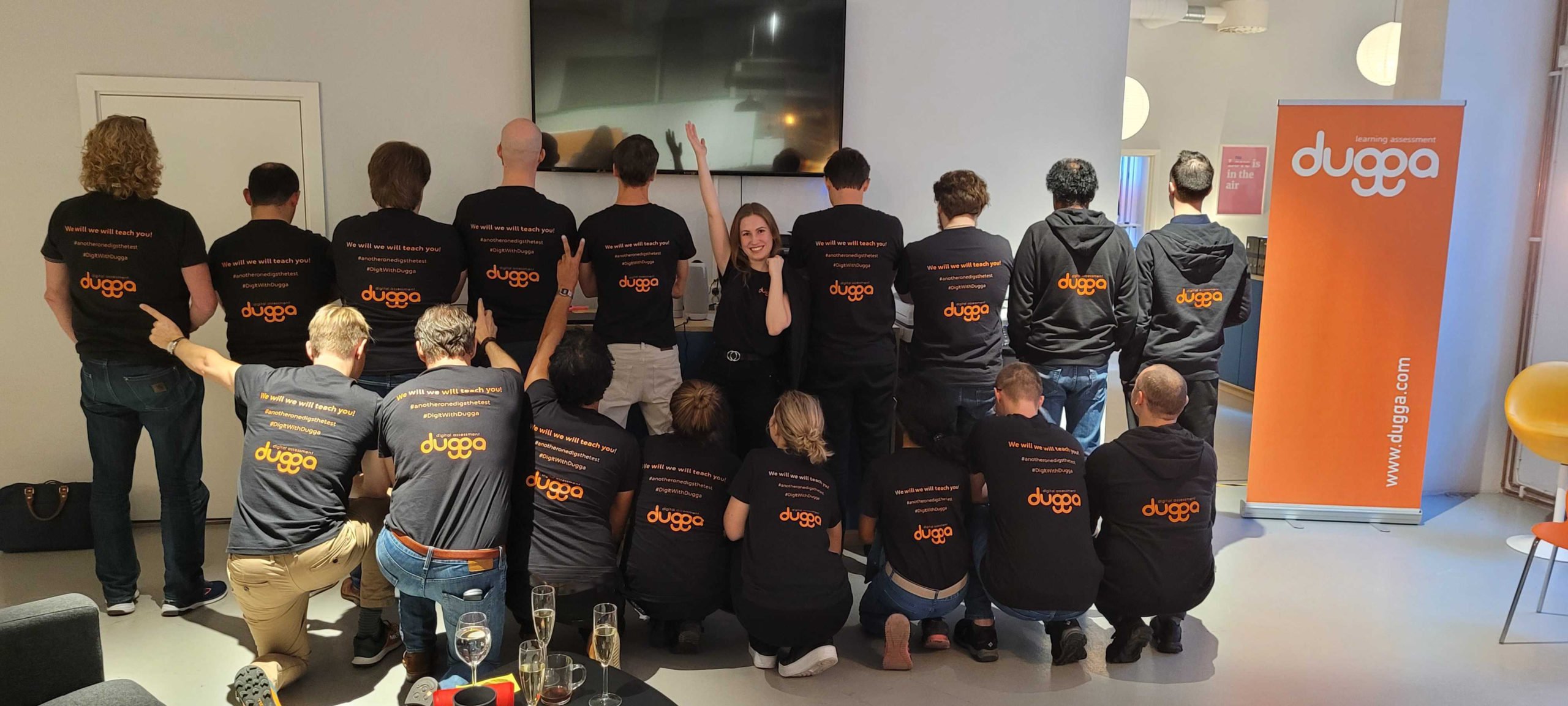 Group in black Dugga shirts, mostly backs visible. One woman smiles & waves. Orange Dugga learning assessment