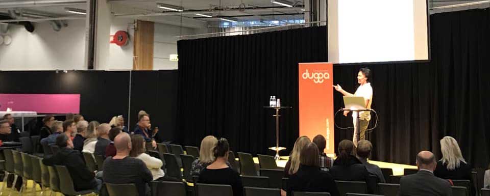 Woman speaker on stage with a "dugga" banner and laptop, presenting to an audience seated in a conference hall.