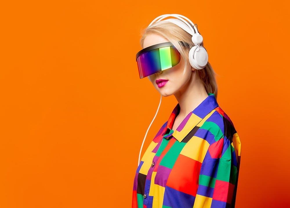 Blonde woman, white headphones, rainbow visor, colorful block shirt, orange background.