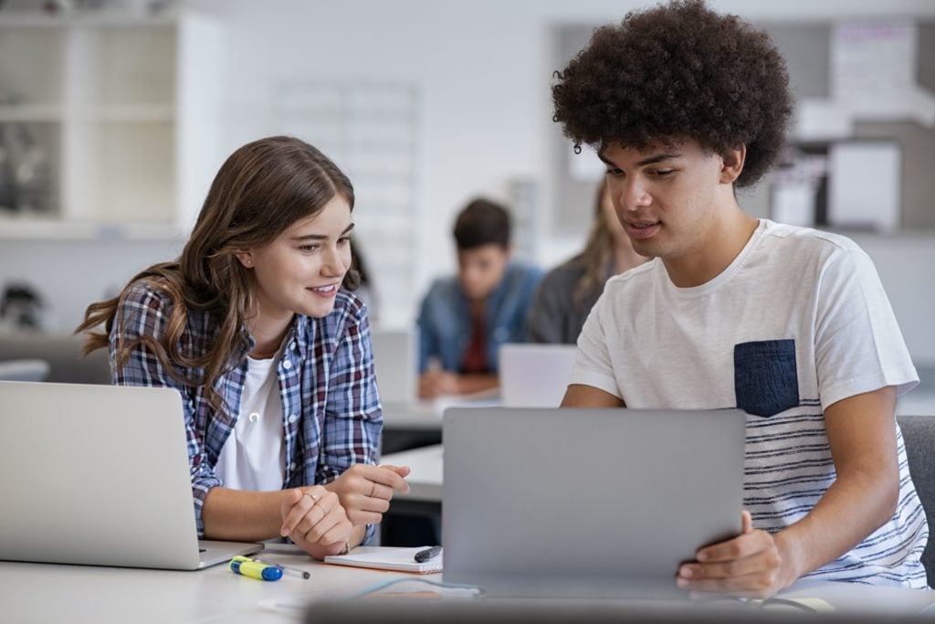 Male and female students look at a laptop in a classroom. Another laptop and stationery are on the desk.