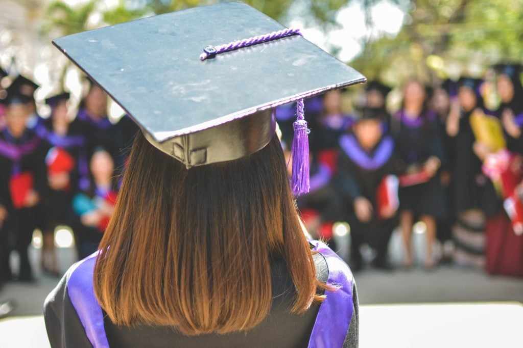Back of a graduate wearing a cap with a purple tassel and stole, facing a blurred crowd of other graduates.