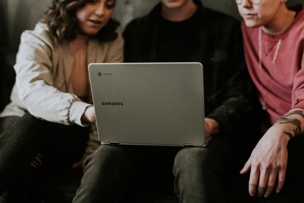 Three people focus on a silver Samsung Chromebook laptop. One points at the screen, another has a tattooed arm.
