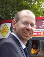 Smiling man in glasses and dark suit. Background: red and yellow signs with foreign script, person in vehicle.