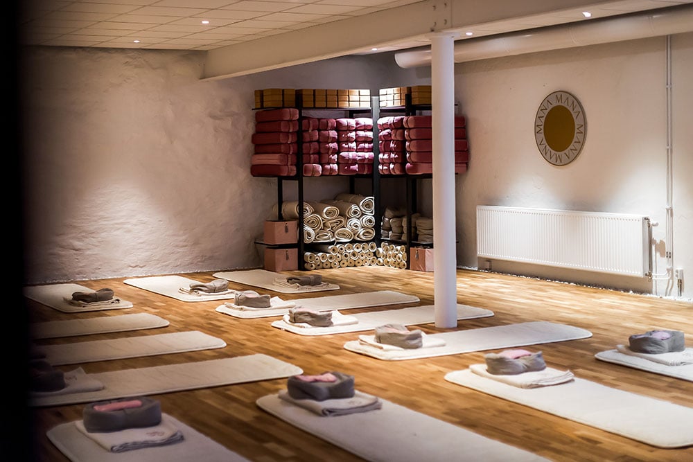 Yoga studio with light wooden floor, white mats with folded towels & small bolsters, shelves of yoga props, and a 'MANA' logo.