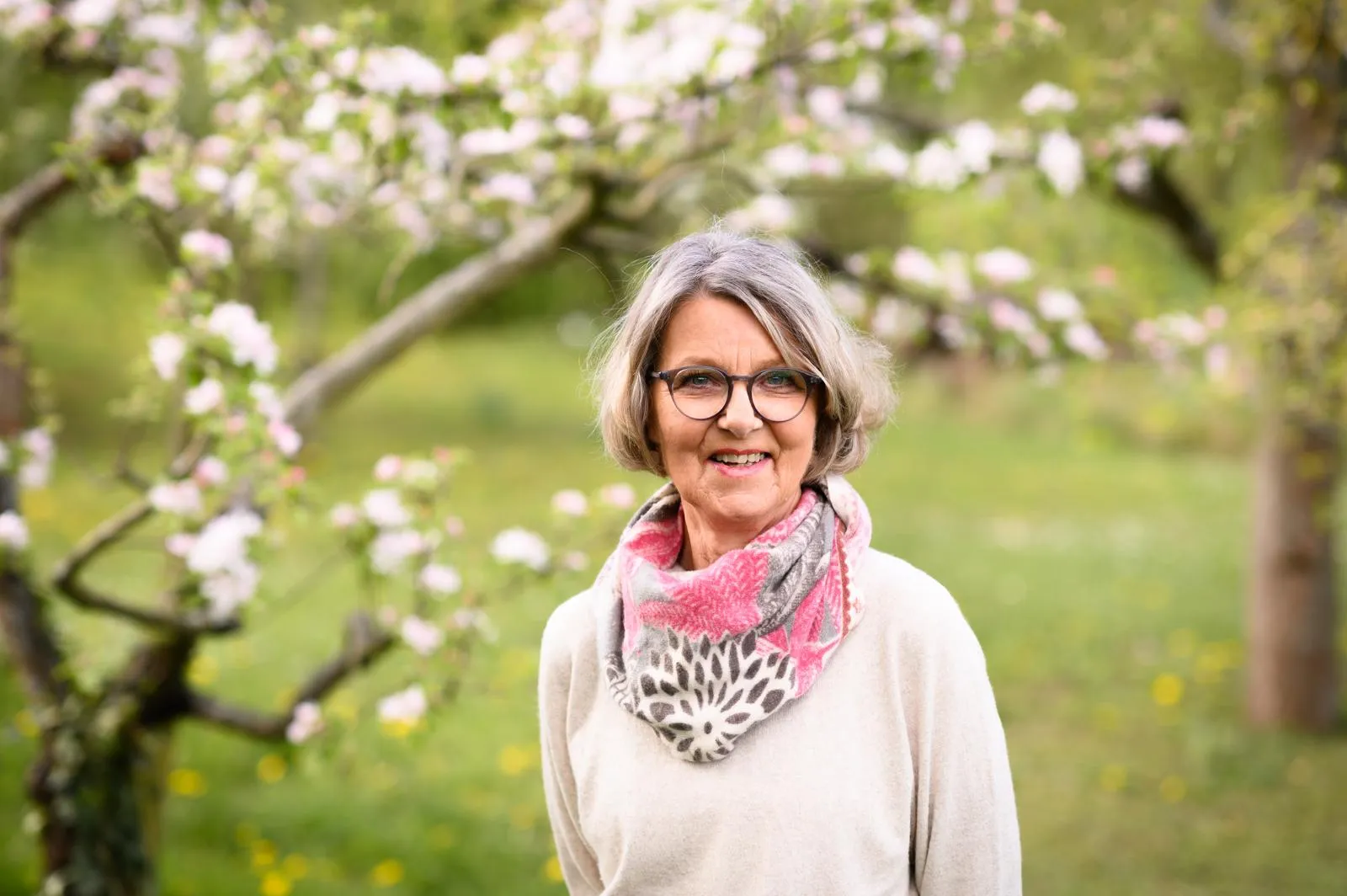Ältere Frau mit grauen Haaren, Brille und buntem Schal steht im Garten mit blühenden Apfelbäumen.