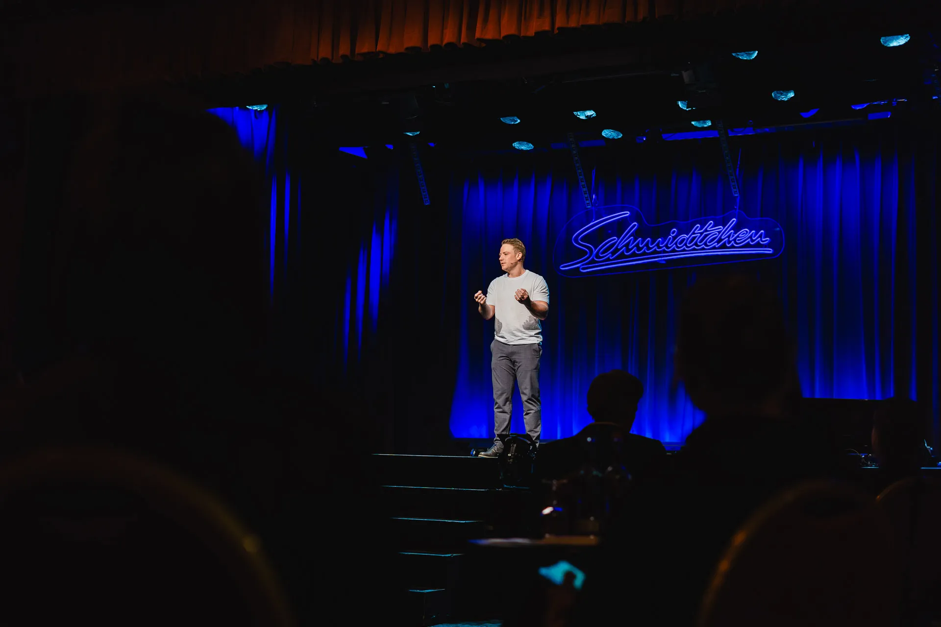 Mann steht auf einer Bühne vor blau beleuchtetem Vorhang und spricht zu einem Publikum, im Hintergrund leuchtet ein Schild mit der Aufschrift „Schmidtchen“.