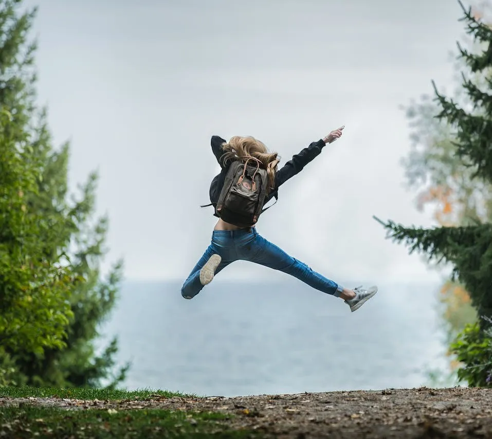 Junge Frau mit Rucksack springt voller Energie in die Luft, im Hintergrund Bäume und ein See.