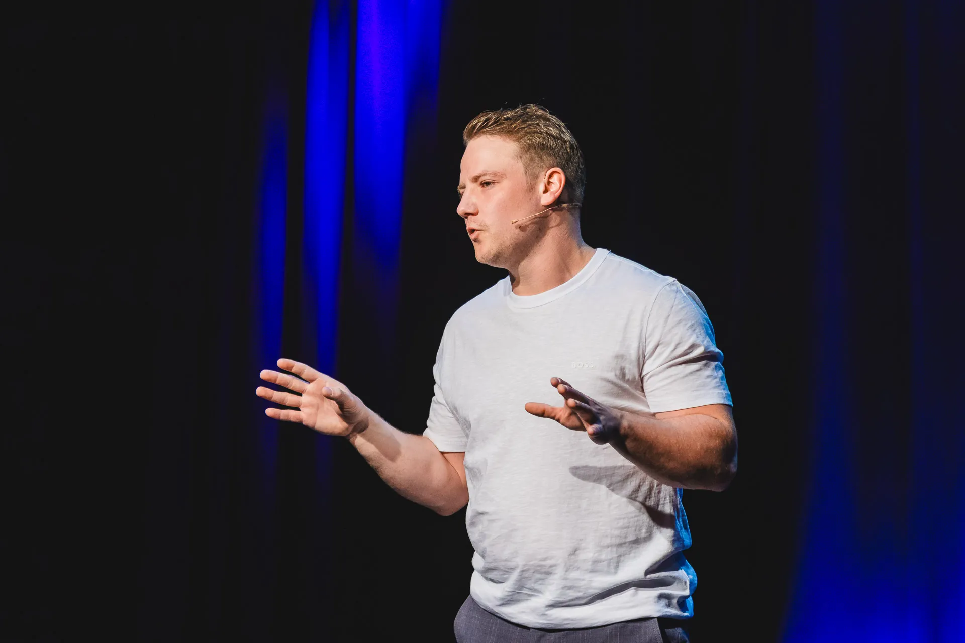 Ein Mann in weißem T-Shirt spricht mit gestikulierenden Händen auf einer Bühne mit blauem Licht im Hintergrund.