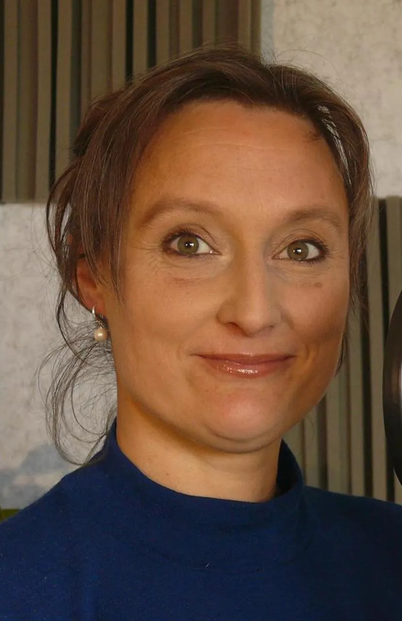 Nahaufnahme einer lächelnden Frau mit braunen Augen und Perlenohrringen, die einen blauen Pullover trägt.