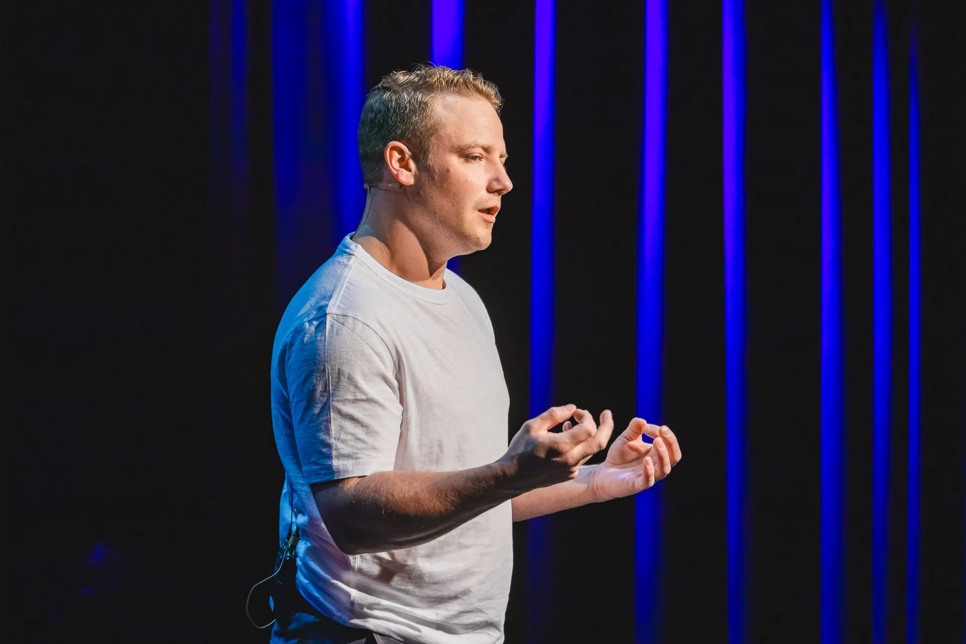 Mann in weißem T-Shirt spricht und gestikuliert auf einer Bühne mit einem Headset-Mikrofon vor einem Hintergrund aus blauen Lichtstreifen.