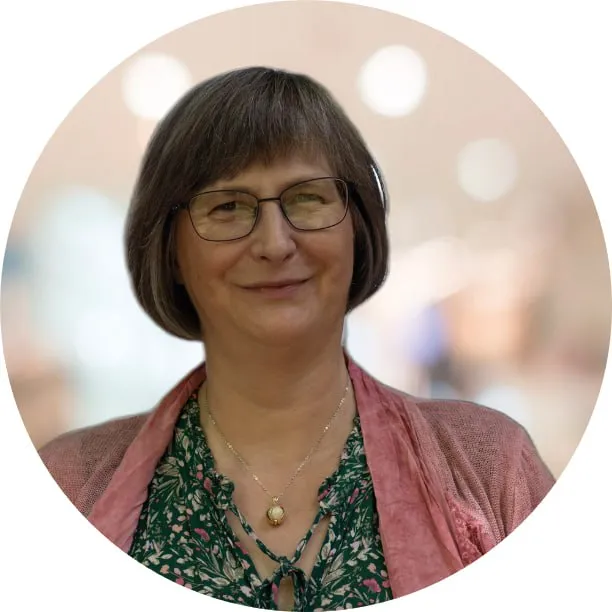 Porträt einer lächelnden Frau mit kurzen braunen Haaren, Brille, grün geblümter Bluse und rosa Cardigan.