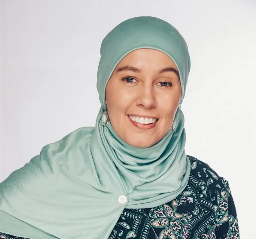 Frau mit hellem Kopftuch und gemustertem Oberteil lächelt vor hellem Hintergrund.
