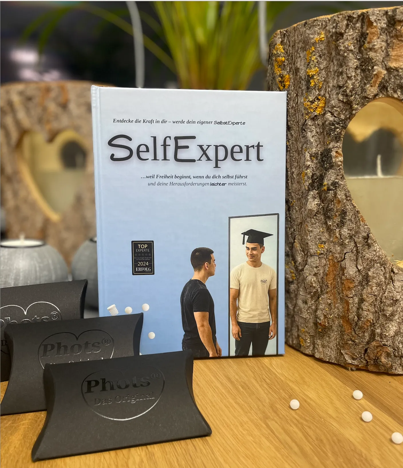 Das Buch 'SelfExpert' steht auf einem Holztisch neben Holzdekoration mit herzförmigem Ausschnitt, dekoriert mit kleinen weißen Kugeln und schwarzen Verpackungen mit dem Logo 'Phots'.