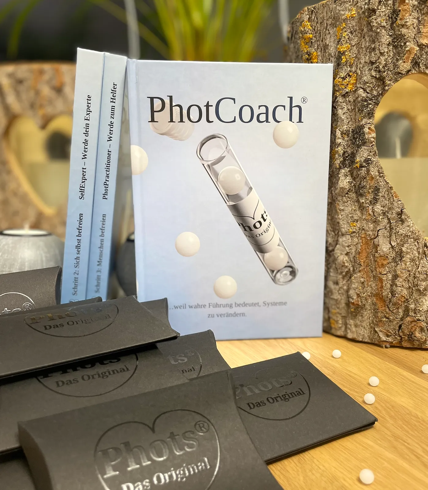 Bücher und schwarze Verpackungen mit der Aufschrift 'PhotCoach' und 'Phots Das Original' auf einem Holztisch, neben einem Baumstamm mit herzförmigem Ausschnitt.